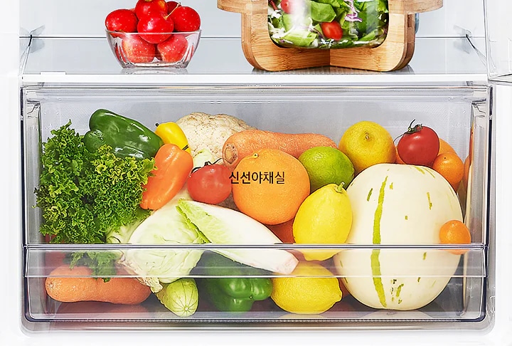 냉장고의 야채 보관실에 다양한 채소와 과일이 가득 차 있습니다. 작은 유리 그릇에 체리가 담겨 있고, 나무 도마 위에는 샐러드가 놓여 있습니다. 야채 보관실 안에는 상추, 피망, 당근, 양배추, 레몬, 오렌지, 멜론 등이 보이며, 신선야채실이라는 글자가 적혀 있습니다.